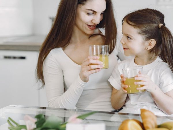 Happy mother and daughter in casual clothes drinking juice and looking at each other while having breakfast together in contemporary kitchen